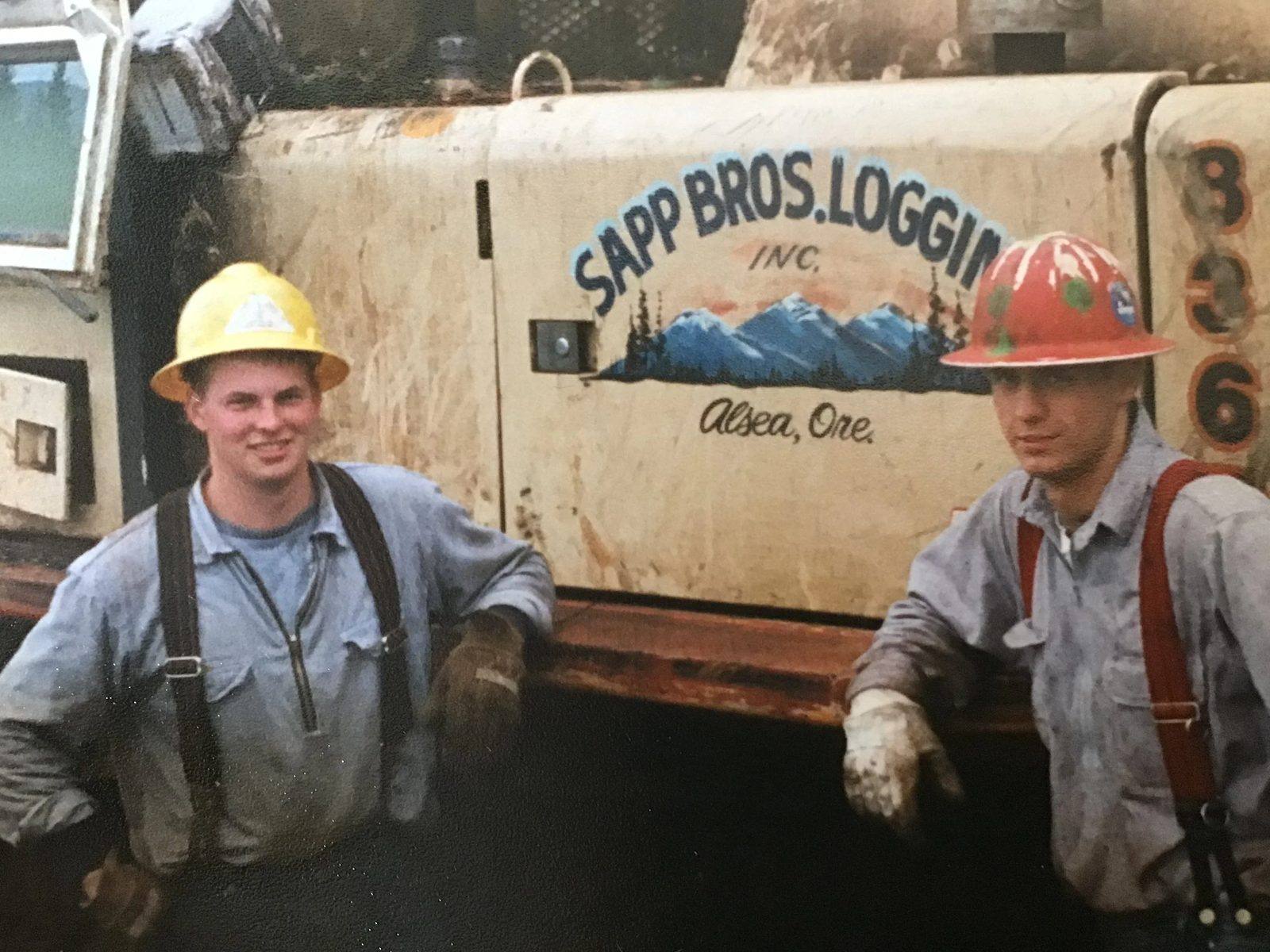 Two loggers standing at the cab of a logging truck on a working forest site.
