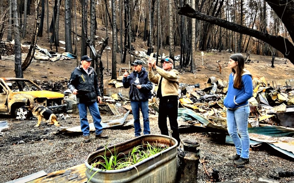 Four people documenting a burned-out property, standing among charred timber, a gutted car and collapsed roofing.