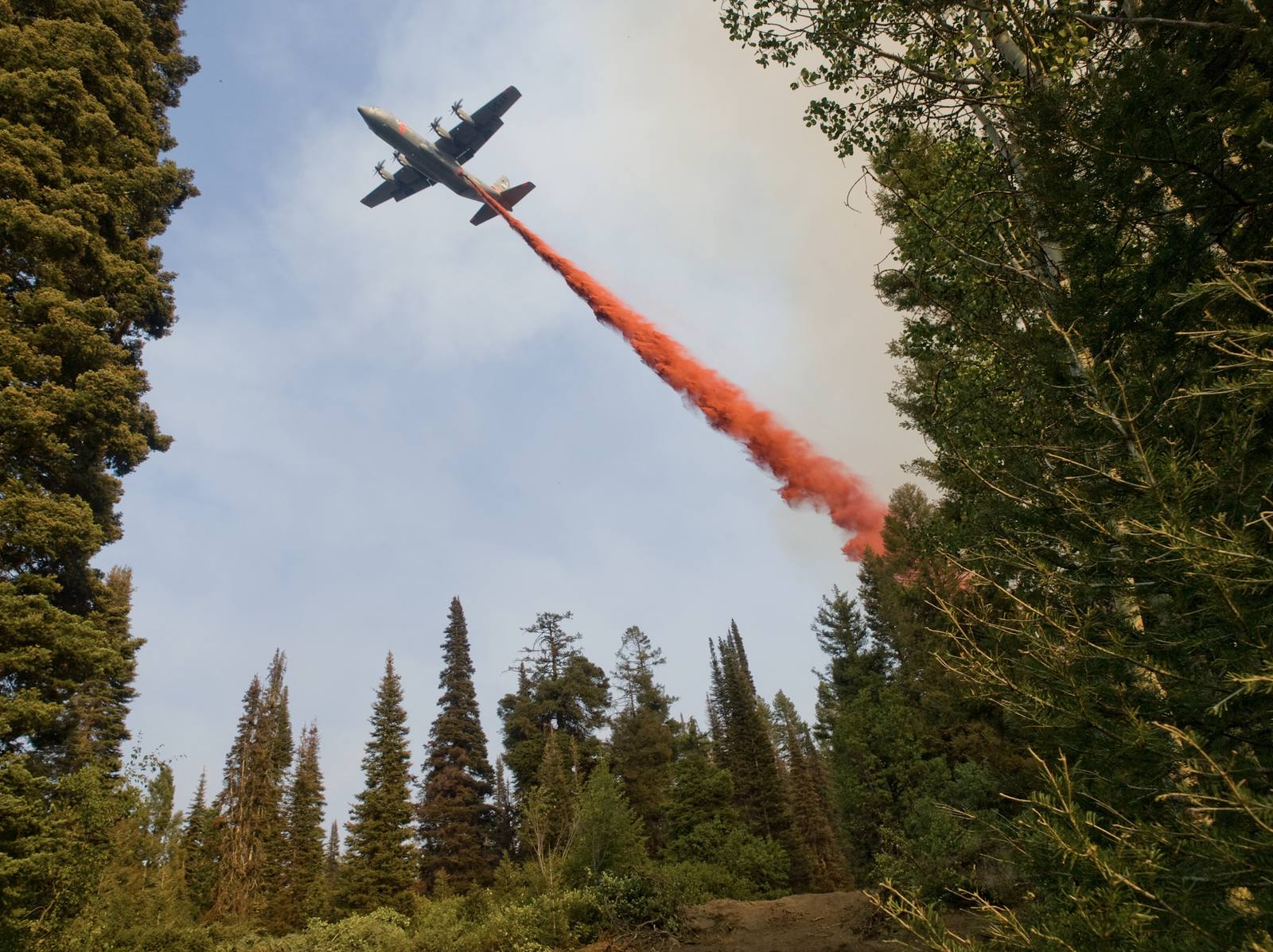 An air tanker banking over a forested ridge as it releases a line of retardant.